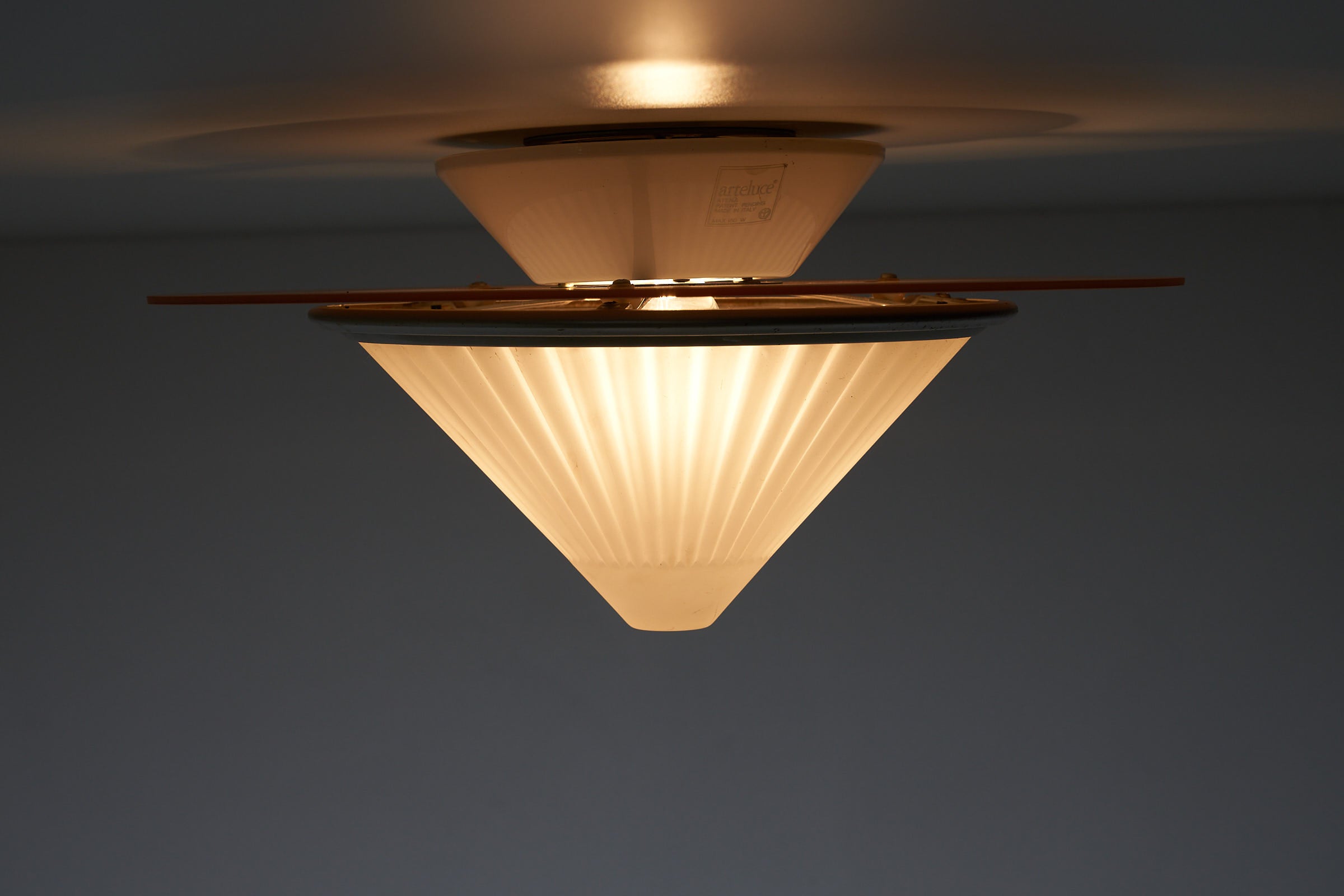 images of the Atena Ceiling Lamp by Ezio Didone for Arteluce, Italy, 1980s. The lamp features a frosted glass cone-shaped diffuser with a colored glass ring around it, separated by a brass ring. The design includes a metal frame that creates a distance between the glass element and the ceiling, producing a backglow effect. The lamp retains its original sticker.