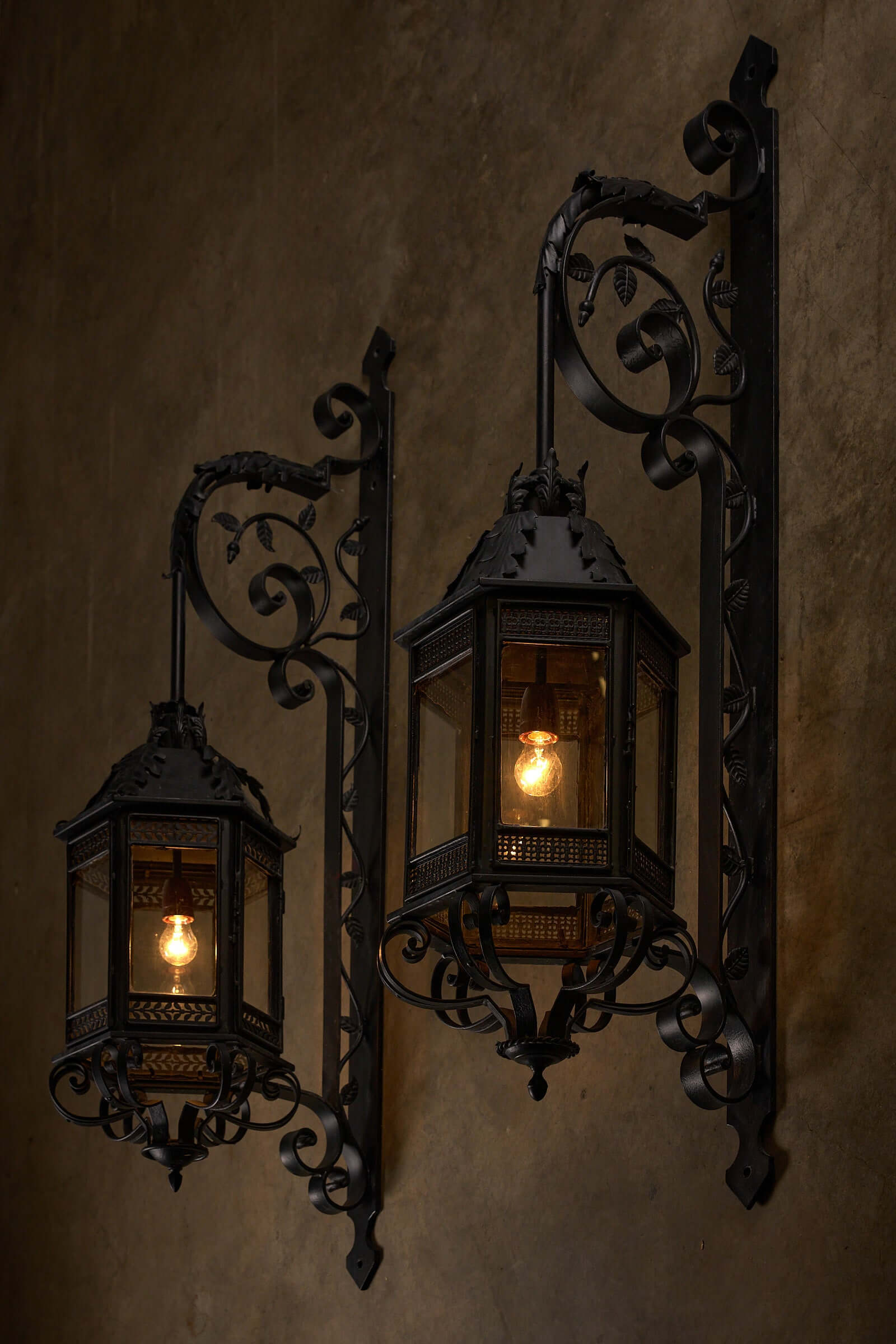 Antique XL wrought iron wall lanterns with intricate scroll designs and glass panels illuminated against a rustic wall.