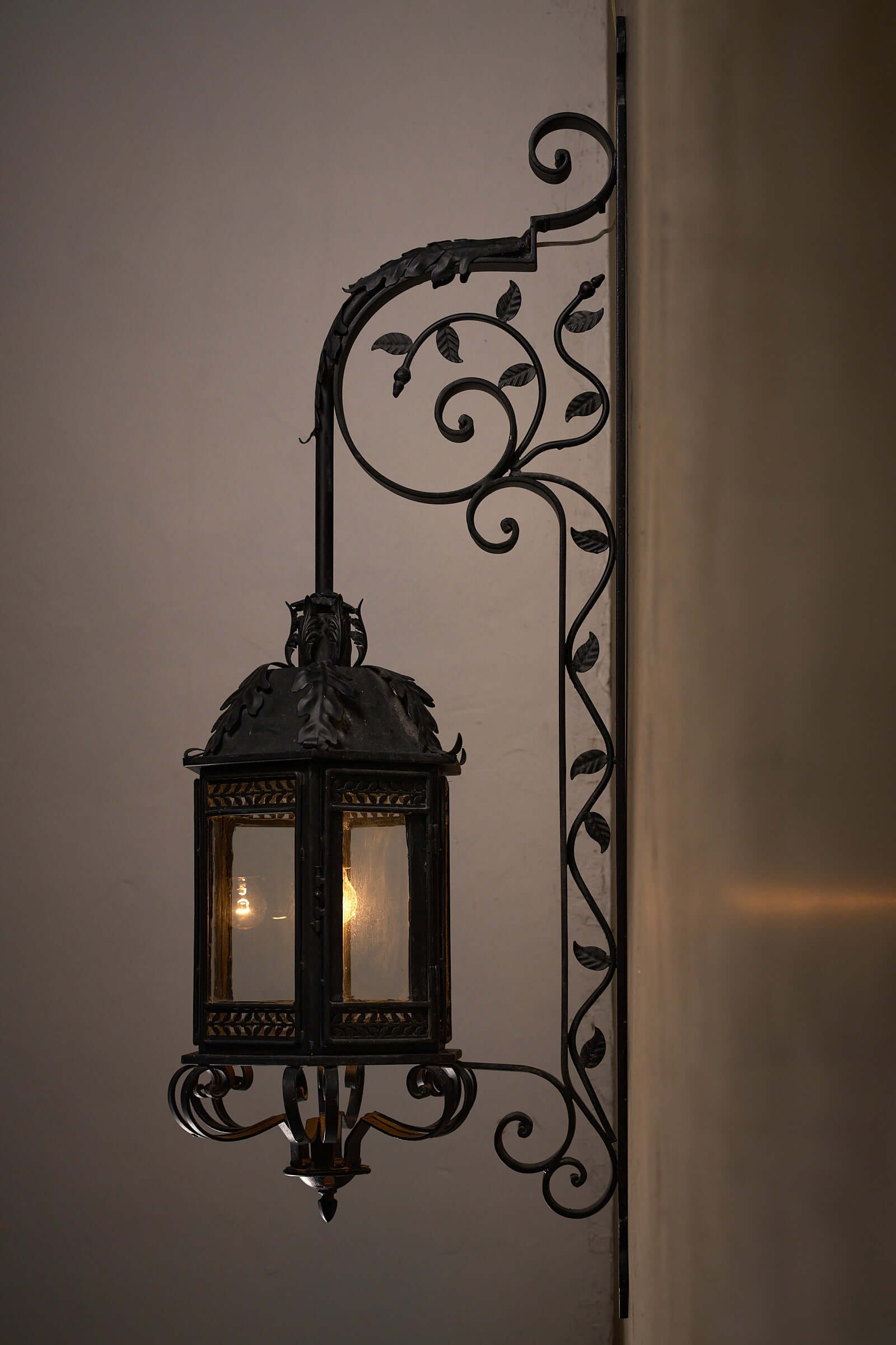 Antique wrought iron wall lantern with intricate scroll designs and glowing glass panels against a neutral background.