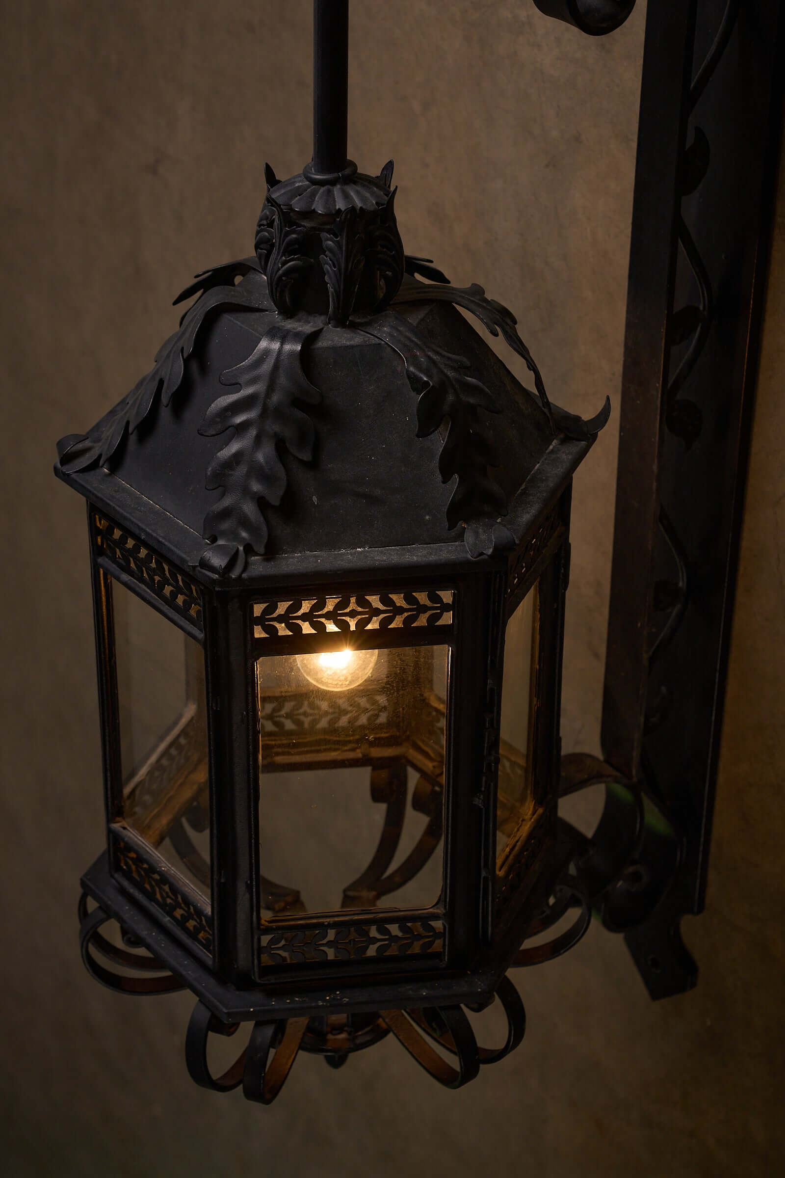 Antique wrought iron wall lantern with intricate scroll designs and lit bulb.