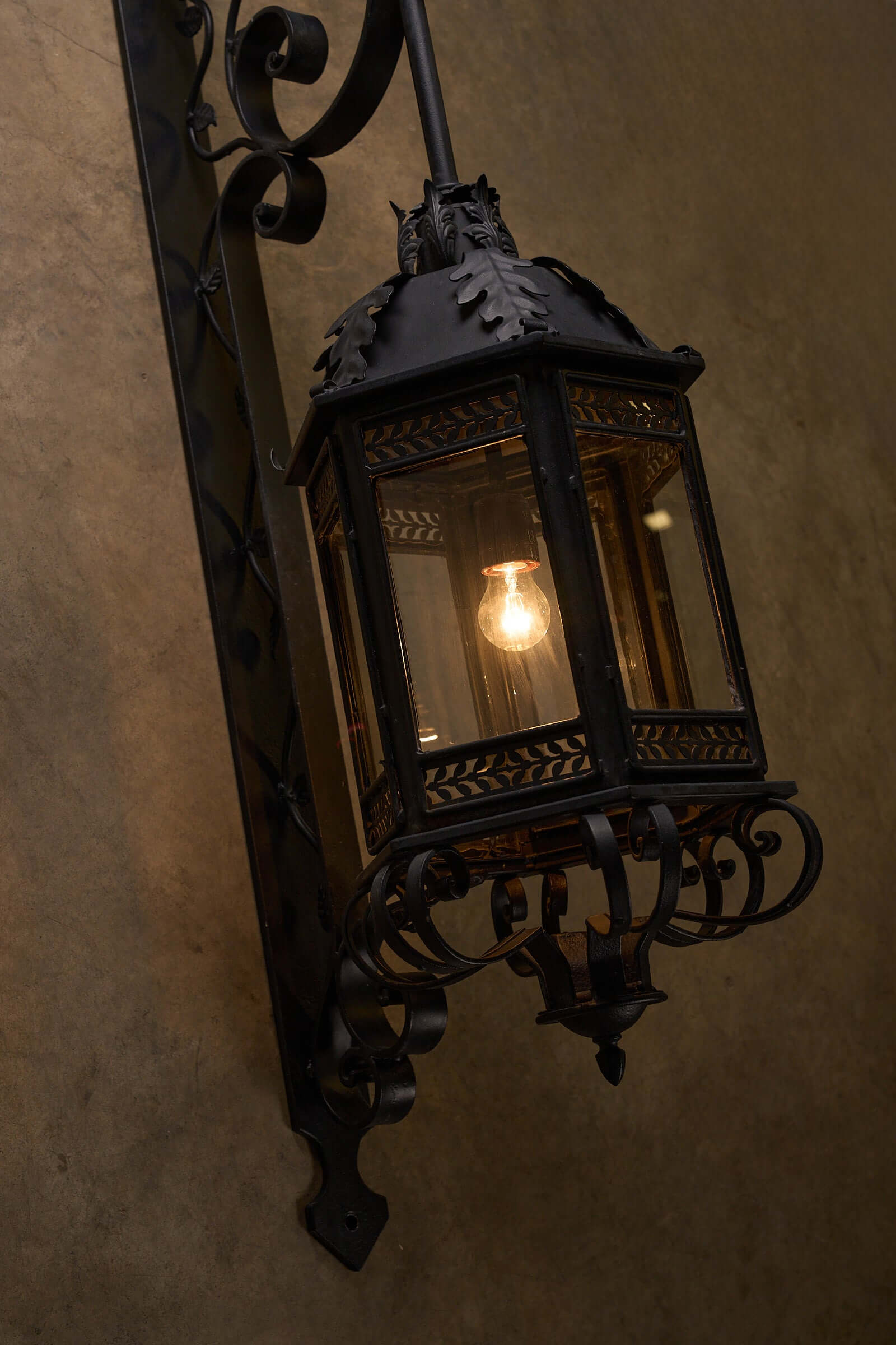 Antique XL wrought iron wall lantern with intricate scroll designs and leaves, mounted on a wall, featuring a glowing light bulb