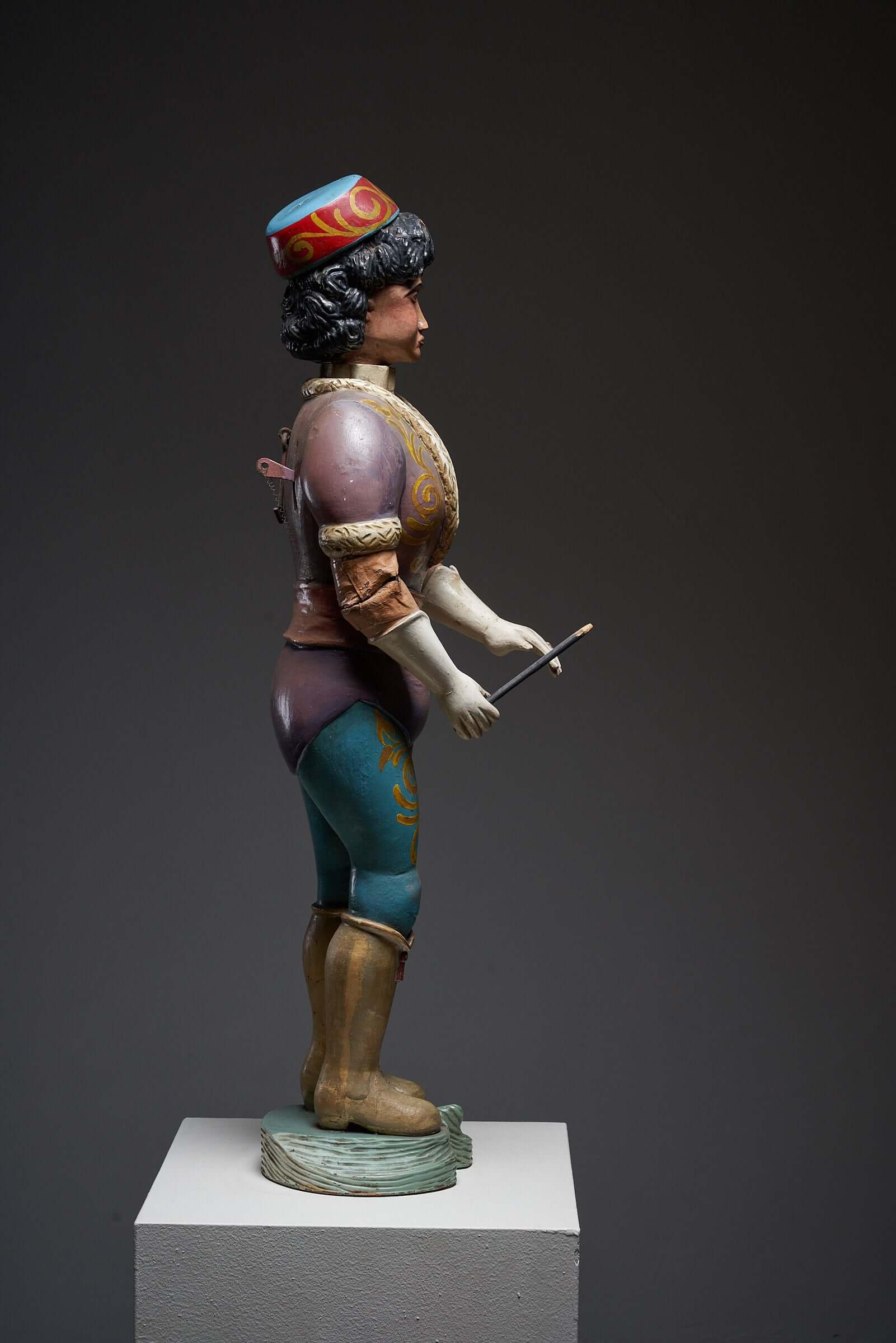 Antique hand-painted wooden carousel conductor figure from the early 20th century in vibrant period attire on a display pedestal