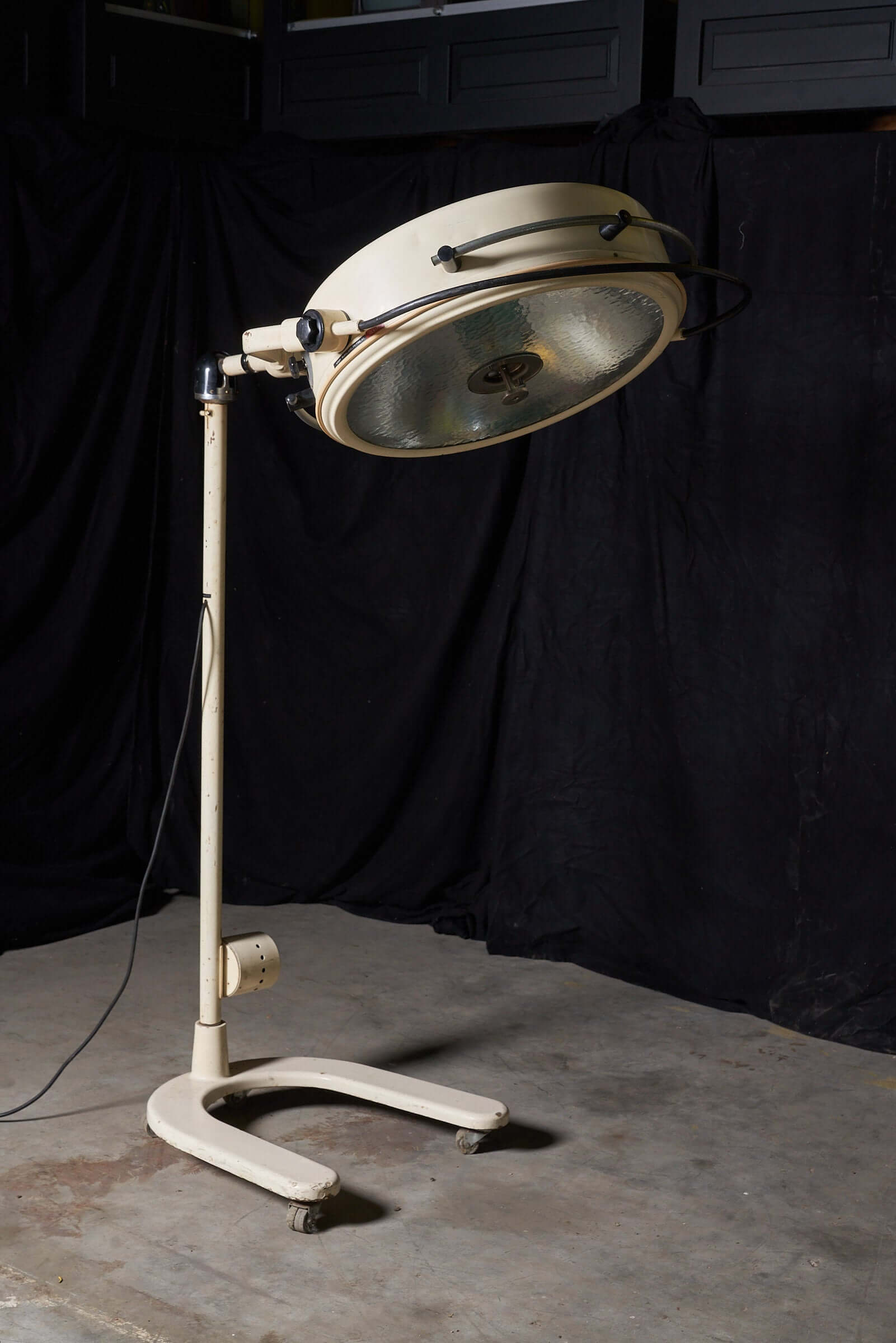 Vintage cream medical lamp with adjustable stand, wheels, and large circular light by Chromophare in an industrial setting.