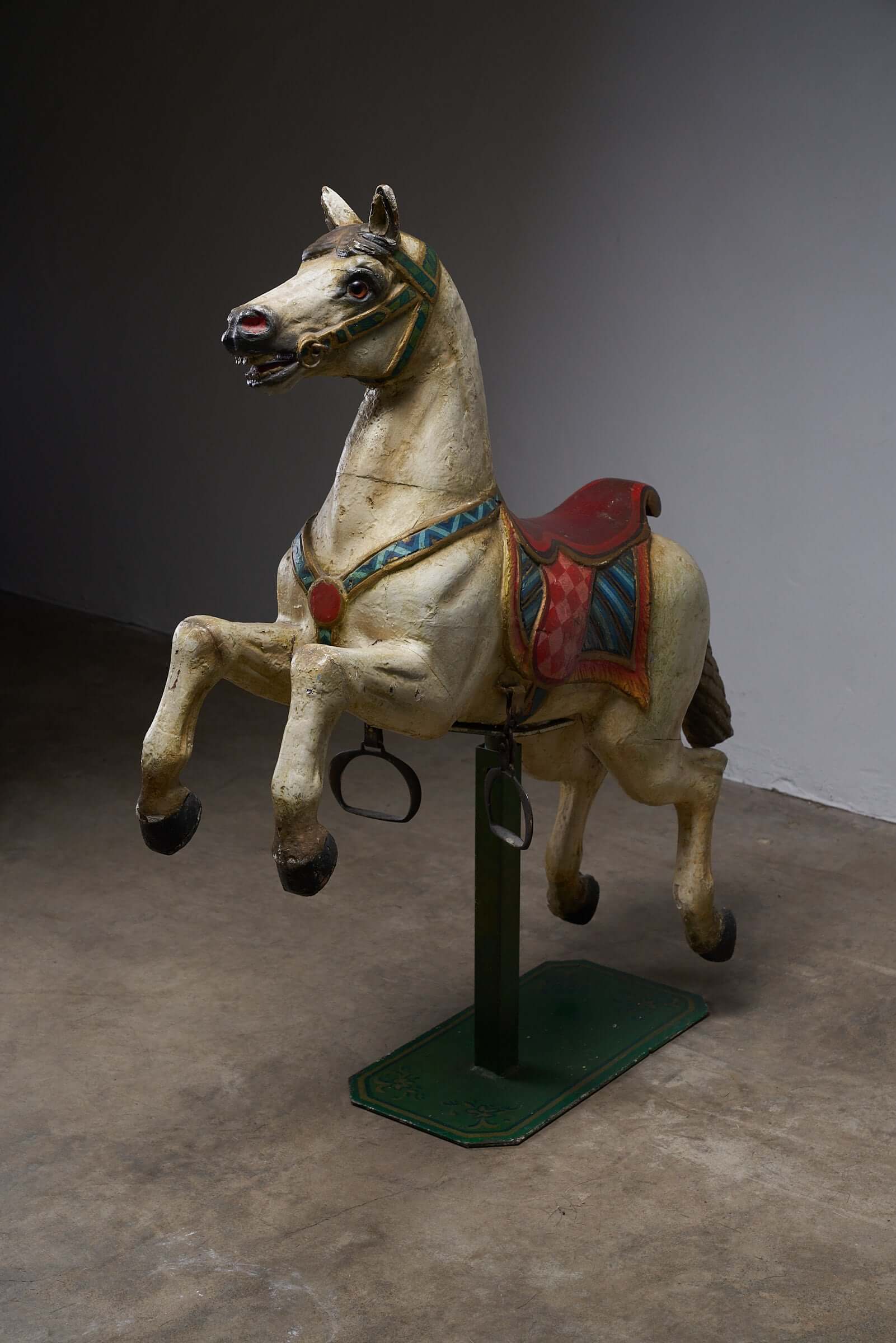 Antique hand-painted wooden carousel horse with multicolored saddle and harness, shown in rearing position on a green stand